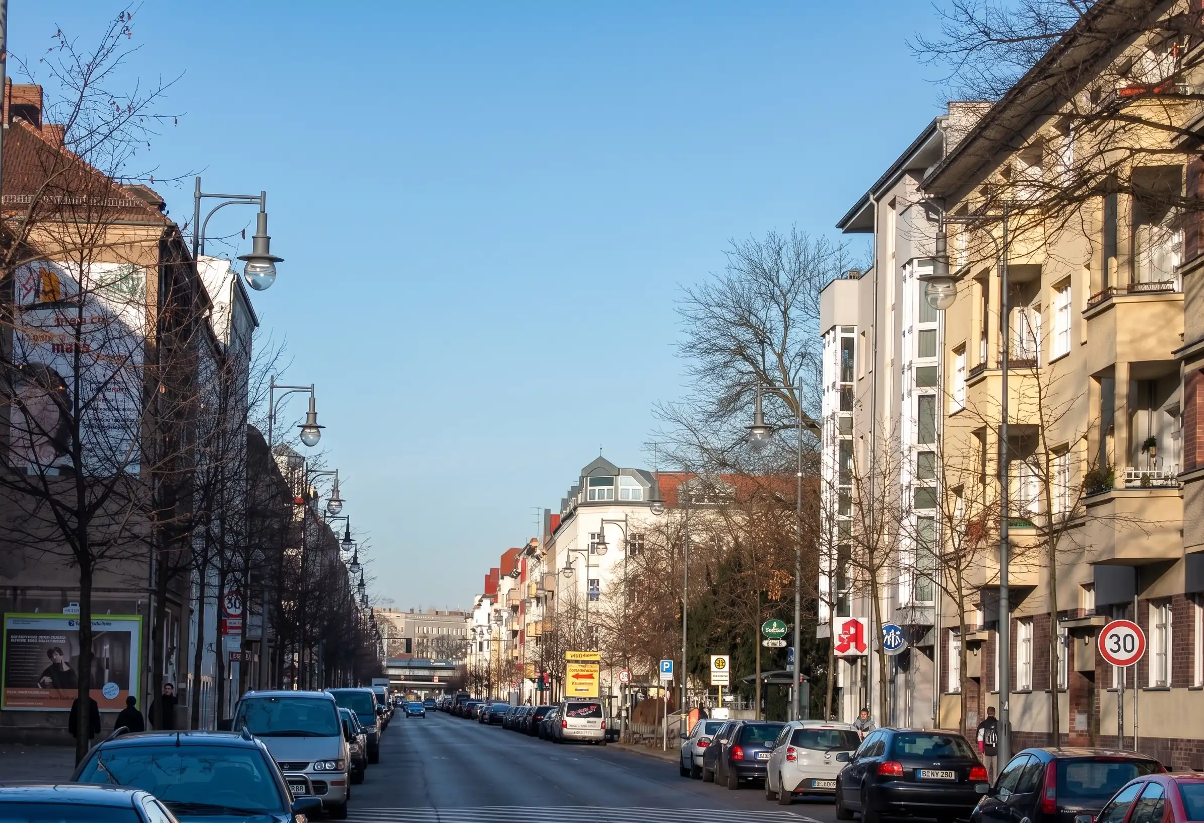 Ausschnitt der Weitlingstraße im Weitlingkiez, Berlin-Lichtenberg, an einem sonnigen Tag. Im Vordergrund eine Apotheke mit dem typischen roten 'A'-Symbol, flankiert von Wohnbauten mit Balkonen und parkenden Autos. Der Blick führt entlang der belebten Straße mit hängenden Laternen unter klarem blauen Himmel.