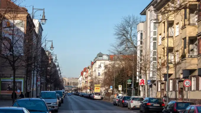 Ausschnitt der Weitlingstraße im Weitlingkiez, Berlin-Lichtenberg, an einem sonnigen Tag. Im Vordergrund eine Apotheke mit dem typischen roten 'A'-Symbol, flankiert von Wohnbauten mit Balkonen und parkenden Autos. Der Blick führt entlang der belebten Straße mit hängenden Laternen unter klarem blauen Himmel.