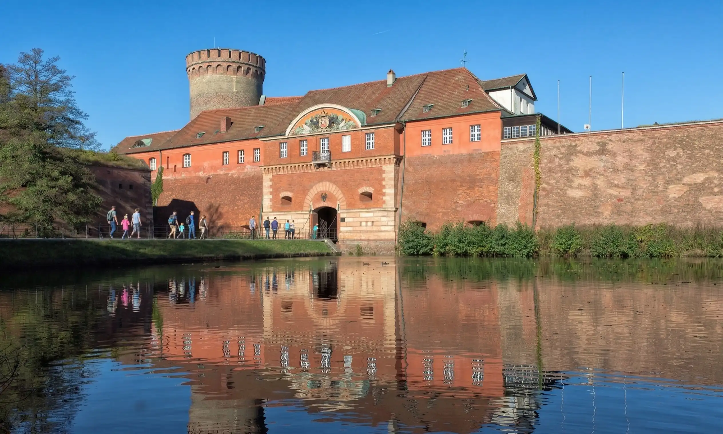 Blick auf die Zitadelle Spandau mit belebter Brücke und Wasserfall-Spiegelung – Ihr Partner für Umzüge im Westen Berlins.